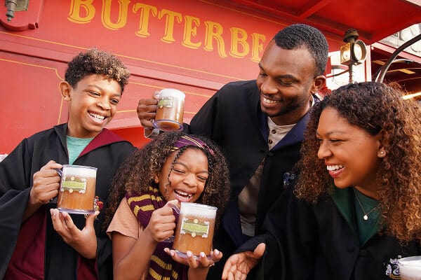 A family dressed in Hogwarts student robes, smiling and enjoying frozen Butterbeer at Hogsmeade.