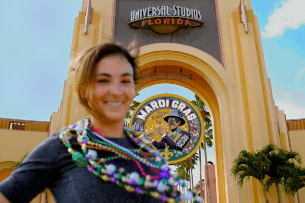 Guests throw beads from atop a parade float at Universal Mardi Gras: International Flavors of Carnaval while a performer dressed like a queen shouts to the crowd.