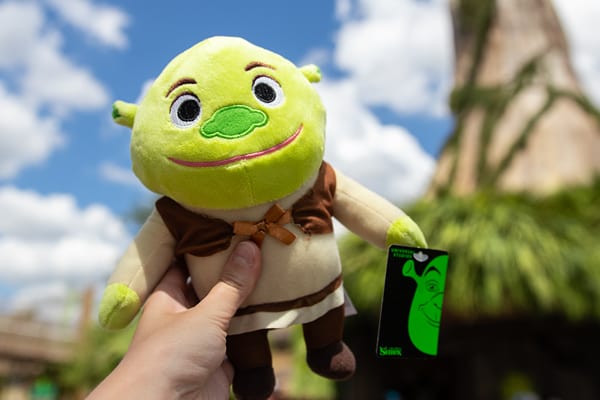 A hand holds up a Shrek plush toy in front of Shrek's cottage in DreamWorks Land at Universal Studios Florida.