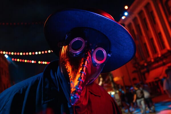 A sinister scareactor with a beak-shaped plague mask, goggles, and black hat bathed in the eerie red light of the night streets.