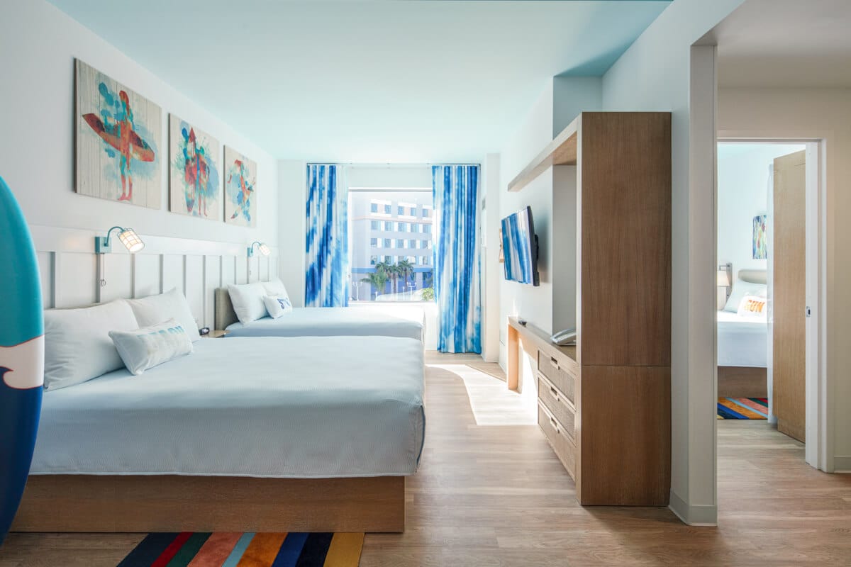 The main room of a two-bedroom suite at Universal Surfside Inn and Suites is decorated with accents of wood and blue and has two queen beds, a dresser, kitchenette, bathroom vanity area and connecting second bedroom.