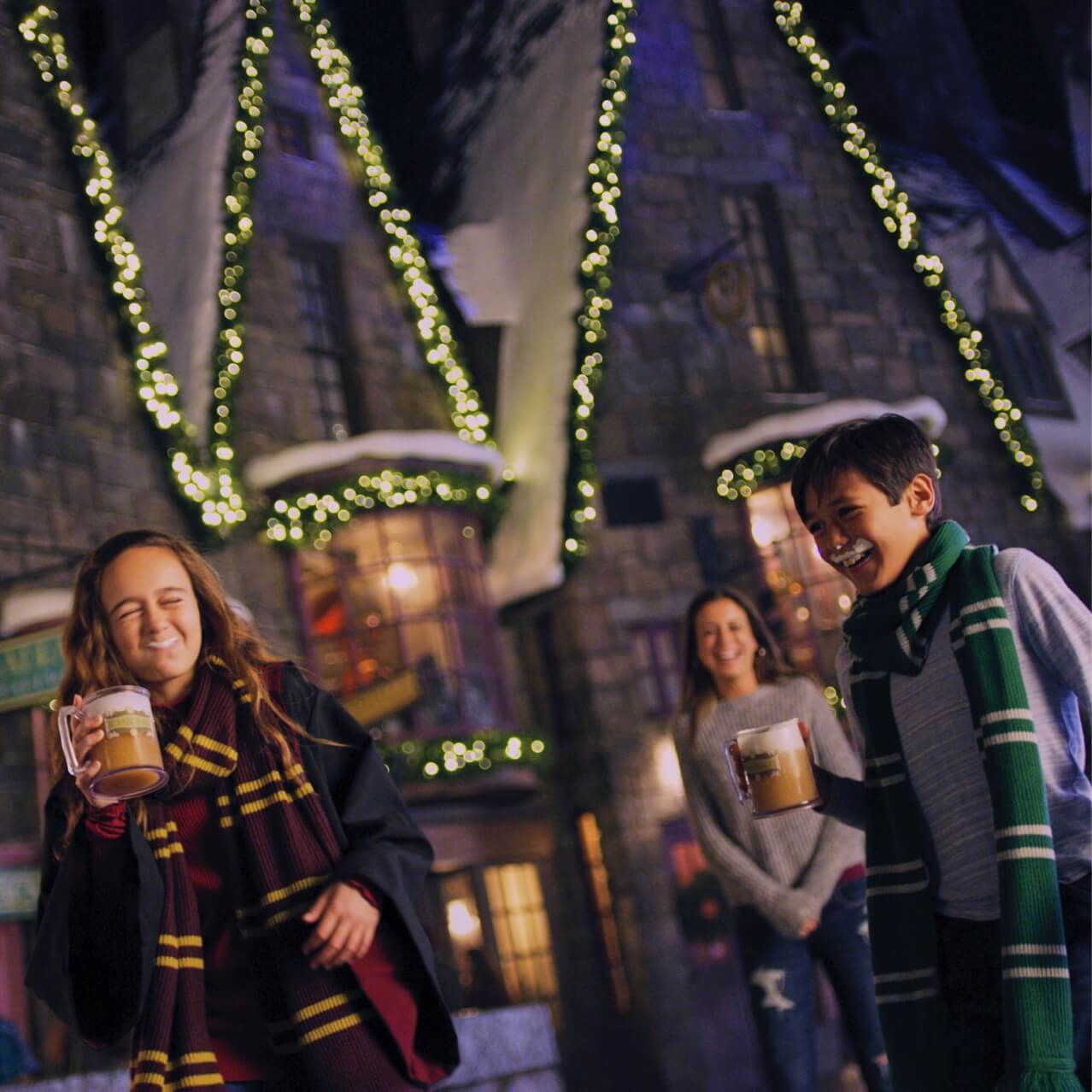 A sister and brother with Butterbeer™ mustaches laugh together in The Wizarding World of Harry Potter – Hogsmeade™, decorated for the holidays.