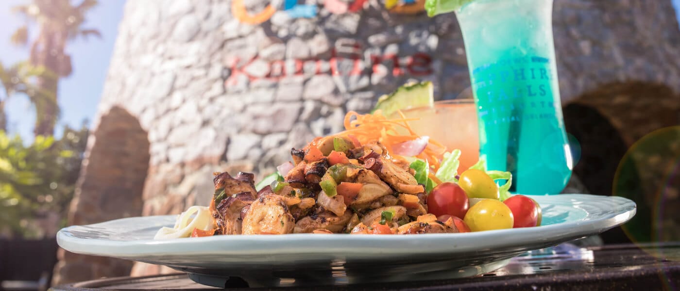 A decorated plated meal and colorful cocktail in front of the Drhum Club Kantine logo sign at Sapphire Falls Resort.