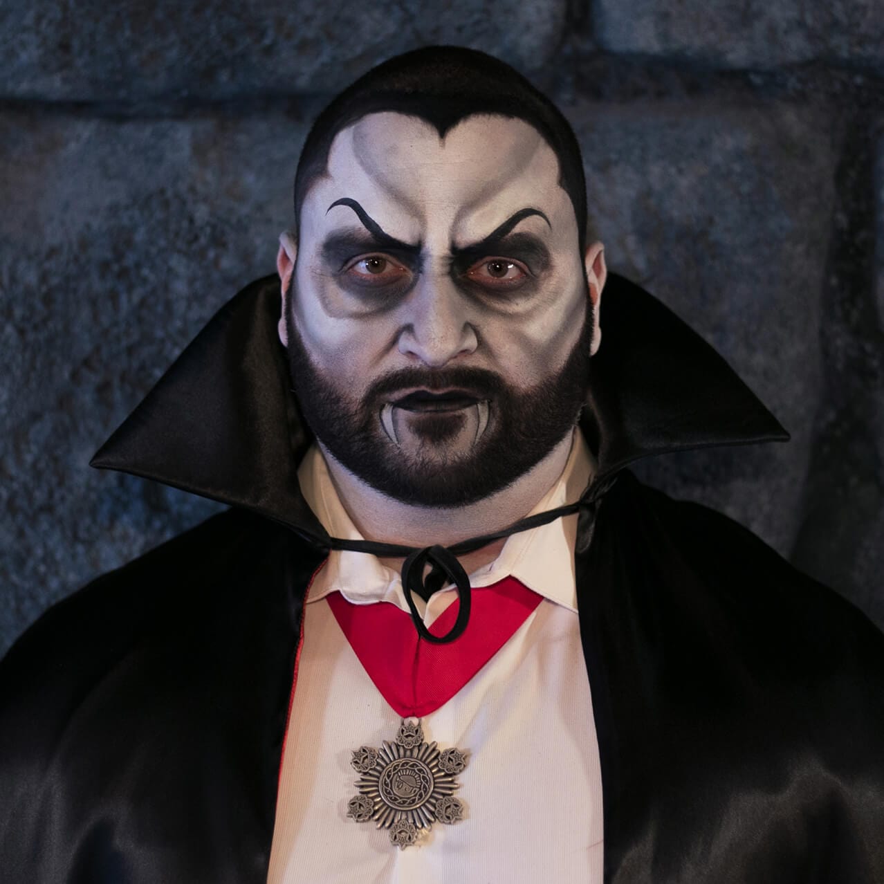 A guest is shown in a before and after shot, smiling on the left and transformed with makeup into a vampire with a cape on the right.