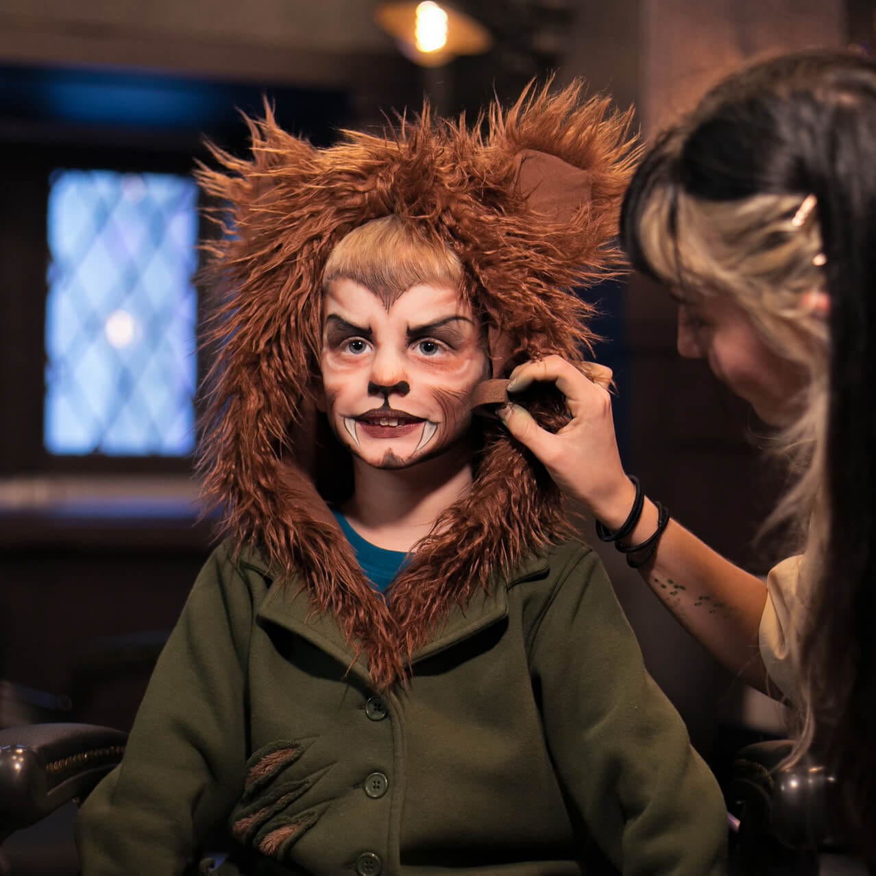 An artist puts the finishing touches on a young customer's face, transforming him into a werewolf in a shaggy hoody at Darkmoor Monster Makeup Experience.