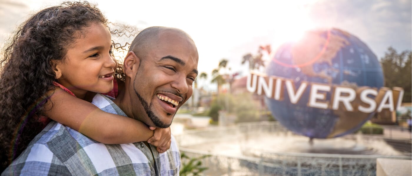 A father laughs as he gives his young daughter a piggy back ride through the park.