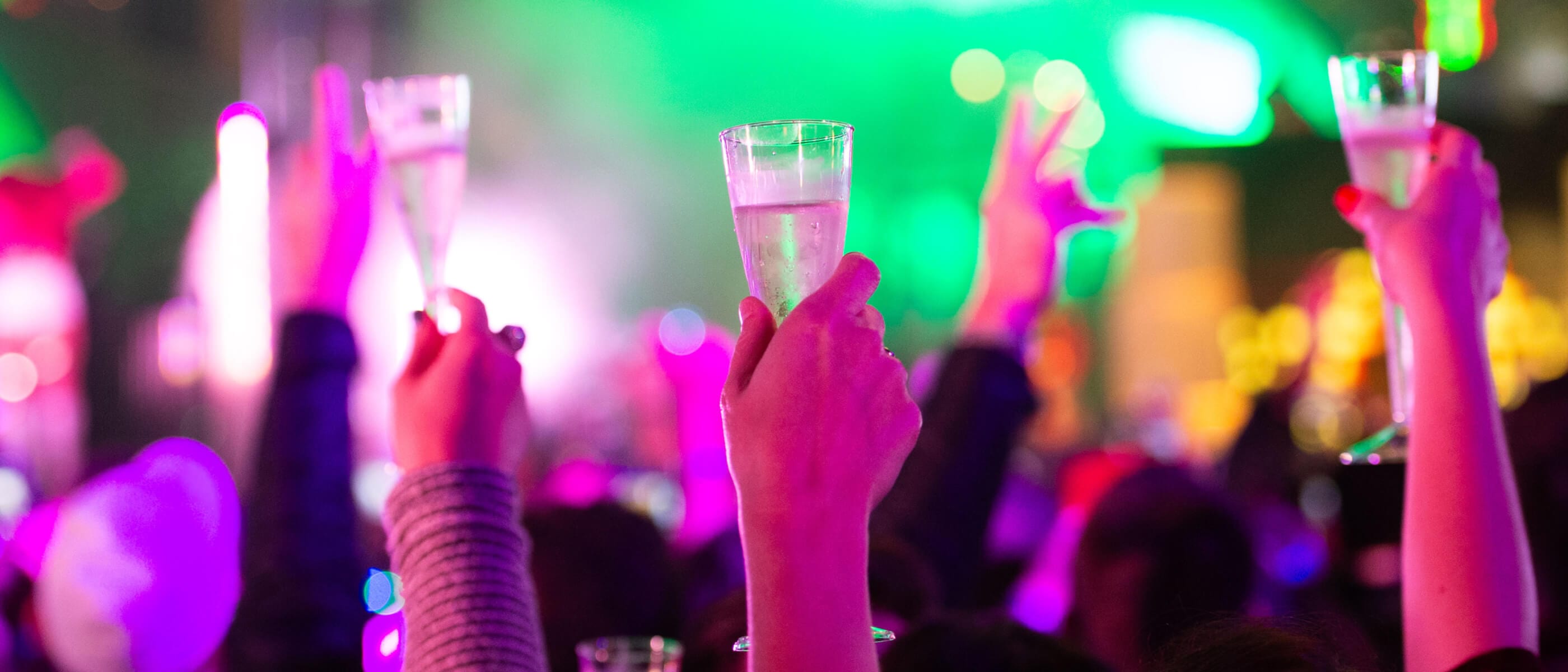 Multiple hands raising their glasses during a champagne toast on New Years Eve with out-of-focus lights in the background.
