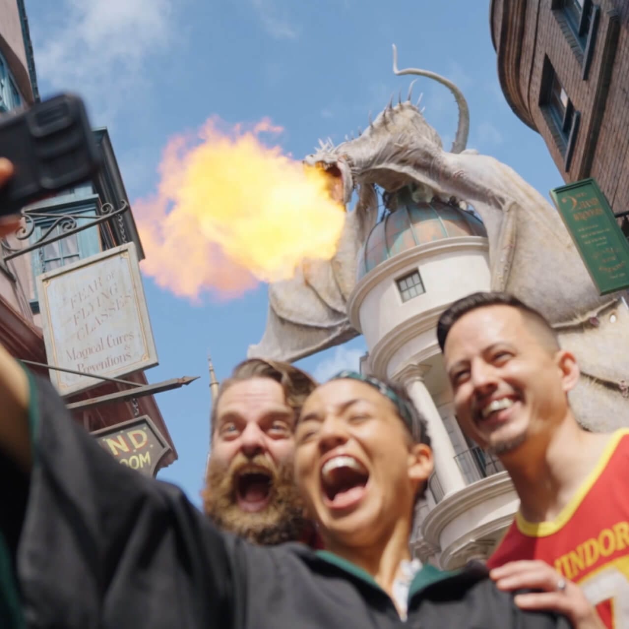 Jimmy, Paige and Jake pose for a selfie in Hogsmeade™ as a dragon breathes flames overhead at Islands of Adventure.
