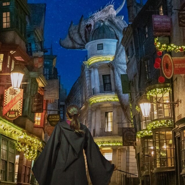 A girl in a wizard robe stands alone in The Wizarding World of Harry Potter™ - Diagon Alley™, which is festively decorated for the holidays.