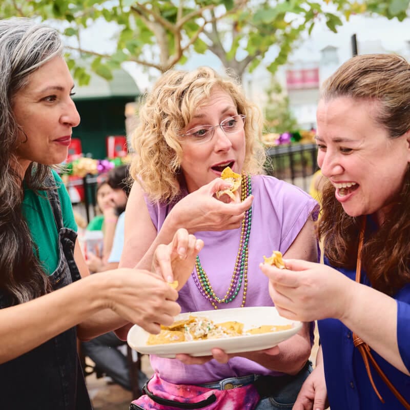 Three friends laugh together as they share a plate of chilaquiles at Universal Mardi Gras: International Flavors of Carnaval.