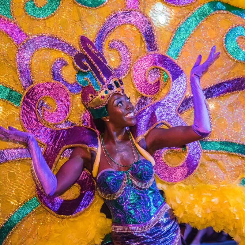 A smiling Mardi Gras performer wearing an elaborate sparkling purple, yellow and teal costume.