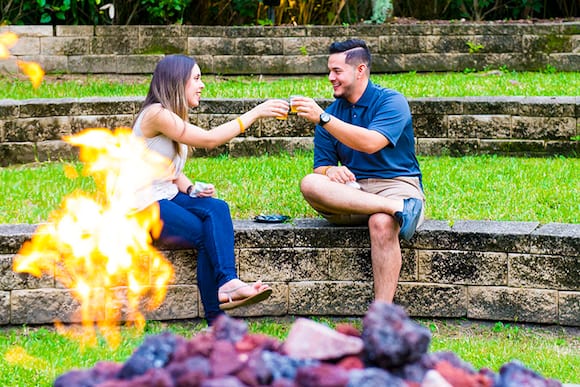 A man and woman toast drinks as they sit near a fire pit.