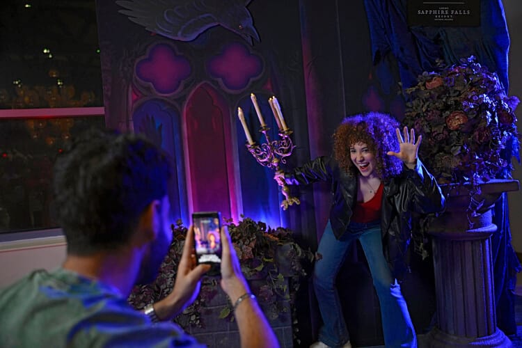 A guest posing with a candelabra in an eerie mansion backdrop while another takes their photo in the Loews Sapphire Falls Resort lobby.