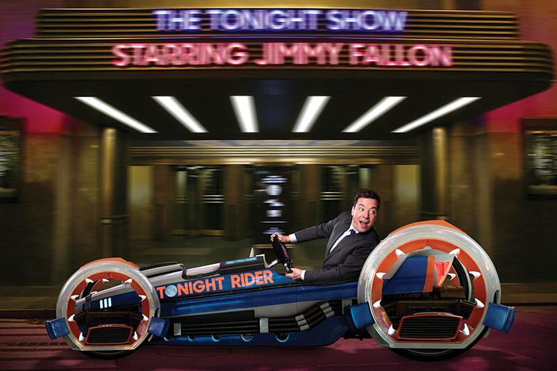 Jimmy Fallon looks back over his shoulder as he speeds along in a blue and red hot rod with the words The Tonight Rider on its side in a scene from the Race Through New York Starring Jimmy Fallon attraction at Universal Orlando.