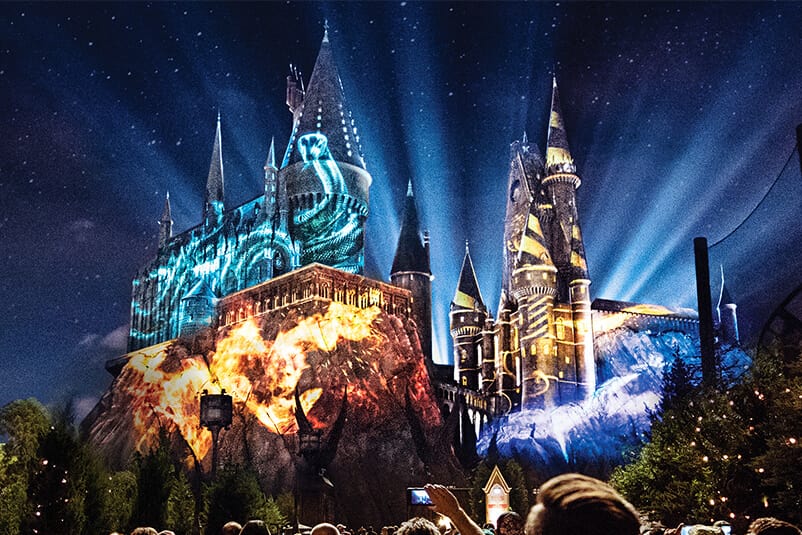 In an artist conceptual rendering, Hogwarts castle is lit up at night with colorful projections depicting the Hogwarts houses.