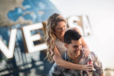 A woman rides on a man's shoulders while holding a Coca-Cola bottle in front of the Universal globe.