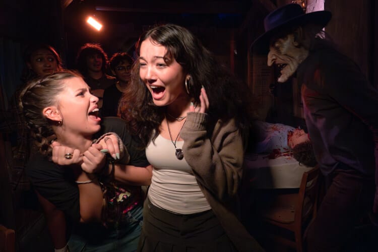 Two screaming friends turning away from a ghoulish scareactor in a black brimmed hat next to a table with a bloody corpse.