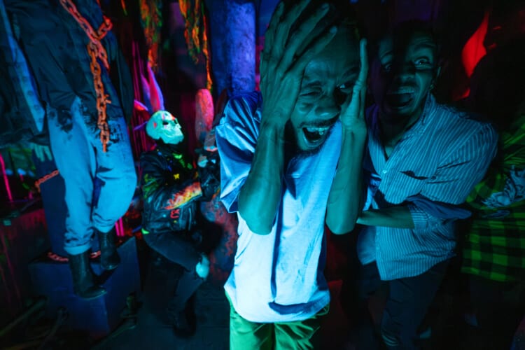 Two haunted house guests screaming in terror as they escape a scareactor with a glow-in-the-dark skull and neon pink mohawk.