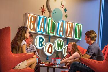 No importa si quieres un snack, un aperitivo, una entrada o una bebida, el restaurante de esta bolera tiene de todo dentro de Cabana Bay Beach Resort. Una familia sentada en una mesa estilo retro disfrutando de una cena cerca del letrero iluminado de Galaxy Bowl en Universal's Cabana Bay Beach Resort.